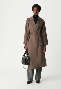 Trench coat in misto lana marrone con vita cintata, ampi risvolti e maniche lunghe, abbinato a pantaloni grigi e una borsa nera.