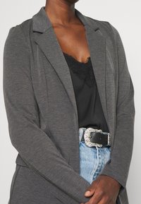 Blazer gris de jersey con cuello de muesca, combinado con una camiseta de encaje negra y denim azul claro. Presenta una hebilla decorativa de cinturón plateada.