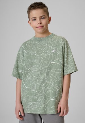 Chłopiec z krótkimi włosami, ubrany w obszerny jasnozielony t-shirt z białym wzorem liści i szare spodnie, stojący na szarym tle.