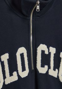 Marinblå zip-up sweatshirt med hög krage, beklädd med elfenbensfärgad fleece-text "POLO CLUB" och en silverfärgad dragkedja. Mjuk, texturerad tyg.