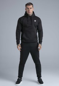 Uomo con tatuaggi che indossa una felpa nera con cappuccio e pantaloni sportivi abbinati, in piedi con le gambe divaricate davanti a uno sfondo grigio uniforme.