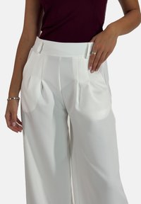 Pantalon blanc à jambes larges avec plis, doté d'une texture lisse et d'une taille cintrée. Le mannequin pose avec une main sur la taille.