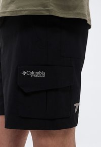 Μαύρα cargo σορτς Columbia Titanium σε άτομο που φοράει ελιά πράσινο πουκάμισο, εμφανίζοντας λεπτομέρειες στον μηρό και στις πλαϊνές τσέπες.