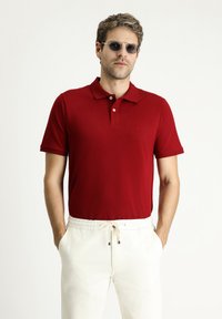 Polo rosso con colletto classico e maniche corte, caratterizzato da tre bottoni e un piccolo logo sul petto. Indossato con pantaloni bianchi a coulisse.