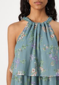 Cvećasti halter top u svijetloplavom materijalu, s naglašenom plisiranoj dizajnu, zelenim listovima i raznobojnim cvjetovima. Lagana tkanina s mekanom teksturom.
