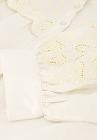 Crème-kleurige blouse met ingewikkelde geborduurde patronen, volants bij de mouwuiteinden en knoopsluitingen aan de voorkant. Zachte, gestructureerde stof.