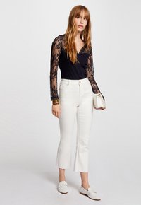 Haut en dentelle noire à manches longues avec un décolleté en V, associé à un jean droit blanc cropped. Des mocassins blancs complètent l'ensemble. Bracelet en or au poignet.