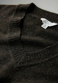 Dunkelbrauner V-Ausschnitt-Pullover mit geripptem Kragen und Saum, aus strukturiertem Wollmaterial. Das Pflegeetikett zeigt die Marke "The Set", Größe M, hergestellt in Bangladesch.