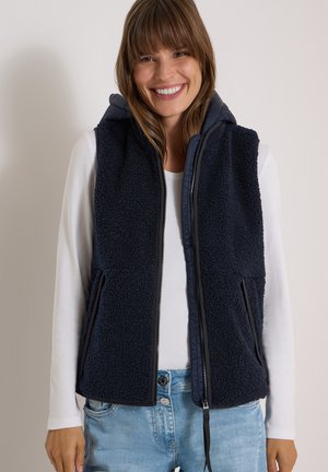 Femme souriante portant une veste polaire bleu marine avec une fermeture éclair par-dessus une chemise blanche à manches longues et un jean bleu clair sur un fond uni.