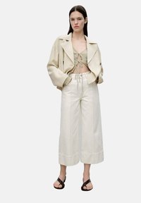 Beige oversize jacket with flared sleeves, paired with a green cropped top and wide-leg white trousers. Black sandals complete the outfit.