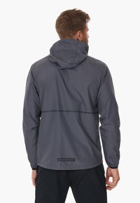 Graue leichte Jacke mit Kapuze, die eine glatte Textur aufweist und einen horizontalen reflektierenden Streifen über den Rücken hat. Branding am Saum angebracht.