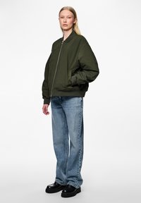 Casaco bomber verde-azeitona com fecho de correr, punhos e barra canelados, combinado com jeans retos de tom azul-claro e sapatos pretos robustos.