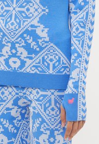 Blue sweater featuring intricate white geometric patterns, ribbed cuffs, and a small pink embroidered accent. Soft knit texture with snug fit.