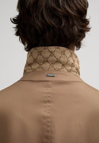 Brauner Blazer mit hohem Kragen; der Kragen ist mit beigem Stoff in braunem Muster versehen. Kleine metallische Logoniete auf der Rückseite.