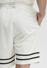 Hvide mesh shorts med en åndbar tekstur, elastisk talje og sorte stribede detaljer nær kanten. Mærket siger "11 PROJECT."
