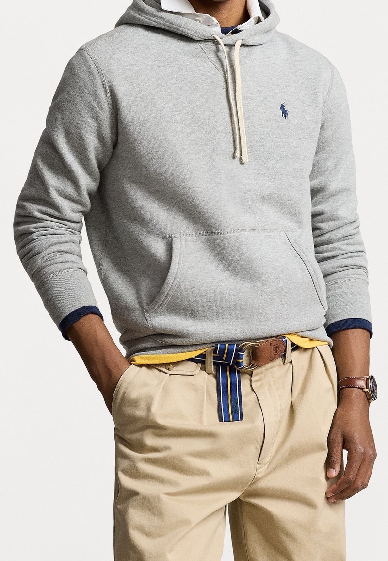 Sudadera gris de algodón con cordón, bolsillo tipo canguro y puños en azul marino. Combinada con pantalones beiges y un cinturón a rayas. Corte casual.