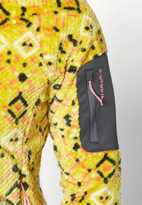 Chaqueta de forro polar amarilla difusa con patrones geométricos en negro y rosa. Presenta un bolsillo en la manga gris con una cremallera rosa y detalle de logo.