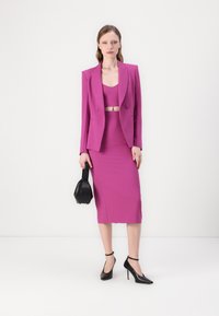 Ensemble fuchsia en deux pièces comprenant un crop top ajusté sans manches et une jupe crayon longueur genoux, associé à des chaussures à talons noirs et à un petit sac.
