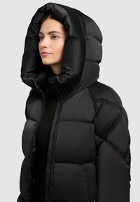 Kvinde iført en sort oversized puffjakke med en stor hætte, lynet halvvejs op, vendt mod venstre mod en neutral baggrund.