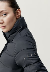 Črna puhovka z visokim ovratnikom, z odlično prešito teksturo in zlatim napisom na levem rokavu. Gladka, sijoča tkanina.