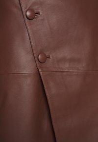 Ruskea nahkavaate, jossa on vinottain oleva läppä, kaksi ruskeaa nappia ja sileä pinta. Saumojen yksityiskohdat näkyvät reunoilla.