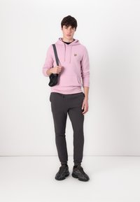Haut à capuche rose clair avec une poche avant et un logo jaune, associé à un jogging gris foncé et des baskets noires. Le modèle tient un sac à bandoulière noir.