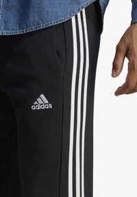 Melni sporta legingi ar baltu trīs svītru dizainu, ar izšūtu Adidas logo kreisajā gurnā un gludu tekstūru.