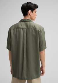 Chemise à manches courtes en tissu vert olive, dotée d'une coupe décontractée, d'un col et d'un détail de pli au dos. Texture lisse sans motifs visibles.