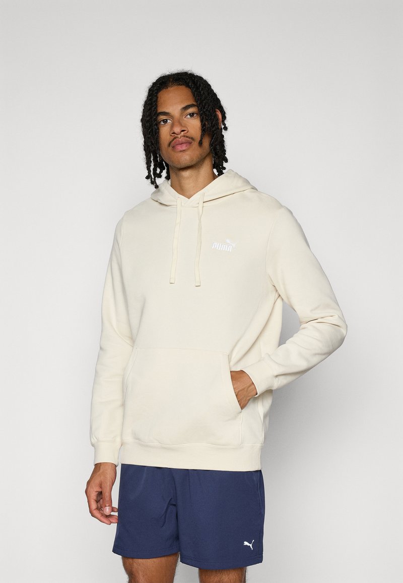 Sweatshirt à capuche couleur crème avec une poche frontale et des cordons de serrage ; présente un petit logo blanc sur la poitrine. Associé à un short marine.