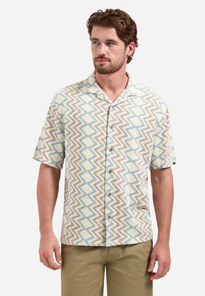 Man draagt een overhemd met korte mouwen met blauw en beige zigzagpatroon, gecombineerd met lichtbruine broek, staand tegen een witte achtergrond.