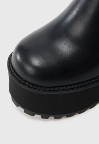 Musta nahkainen nilkkuri, jossa pyöristetty kärki, paksu korokepohja ja ommellut yksityiskohdat reunalla. Kuvioitu kumipohja tarjoaa hyvän pidon.