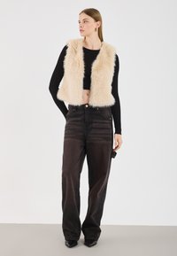 Krämfärgad fuskpäls utan ärmar över en svart långärmad crop top, kombinerat med högmidjade mörka jeans och spetsiga svarta skor.