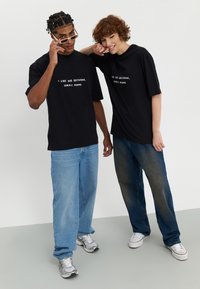 Two individuals wear oversized black t-shirts with white text. The man pairs it with light blue denim jeans and sneakers, while the woman has dark jeans.