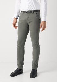Pantaloni in cotone slim-fit di colore verde oliva, con tasche frontali, una cintura nera e abbinati a mocassini neri, su uno sfondo semplice.