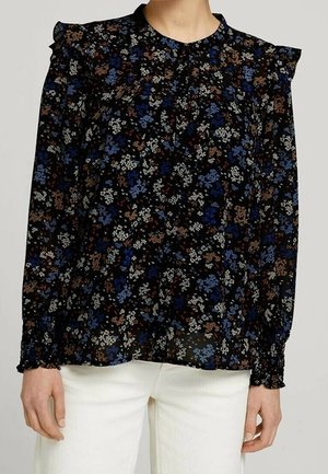 Femme portant un chemisier noir à manches longues avec de petits imprimés floraux bleus, blancs et marron et des détails à volants aux épaules, associé à un pantalon blanc.