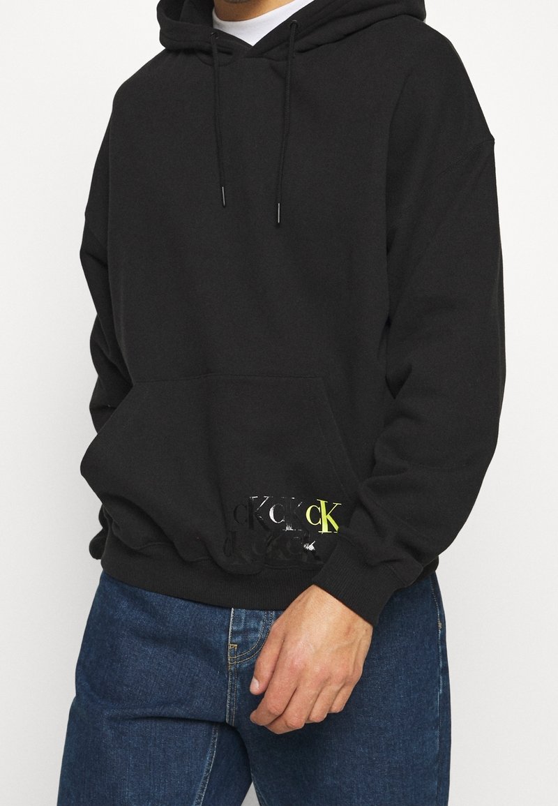 Sudadera negra de tela de mezcla de algodón. Presenta un bolsillo frontal, cordones y un logo en negro brillante y amarillo neón en el dobladillo.