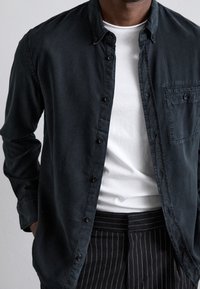 Camicia nera a maniche lunghe realizzata in tessuto morbido, con tasca frontale e colletto classico, indossata sopra una t-shirt bianca a girocollo.