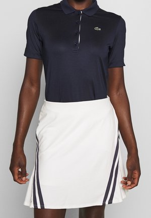 Persona con una camiseta polo negra de manga corta con botones y una falda blanca con dos franjas verticales negras a cada lado.