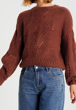Femme portant un pull marron en maille avec un motif de feuilles et des manches bouffantes, associé à un jean bleu taille haute.