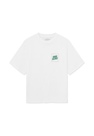 Mand iført brunternet skjorte under en hvid t-shirt med teksten "RADIO SILENCE", sorte bukser og sorte støvler, stående mod en hvid baggrund.