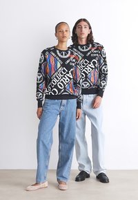 Svart hettegenser med fargerike grafiske trykk og tekst, kombinert med lyseblå jeans. Modellens fottøy inkluderer lyse flate sko og svarte sko.