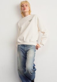 Persoon in een wit sweatshirt en een losse, lichtblauwe spijkerbroek, staand met één hand op de heup tegen een eenvoudige witte achtergrond.