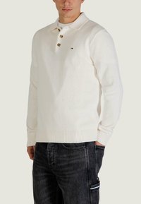 Biały bawełniany sweter polo z długim rękawem, z prążkowanym wykończeniem, trzema brązowymi guzikami i małym logo na piersi. Noszony z ciemnymi dżinsami.