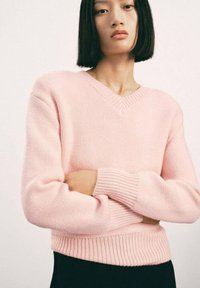 Giovane donna con capelli neri lisci indossa un maglione rosa chiaro a V, incrociando le braccia su uno sfondo chiaro e semplice.