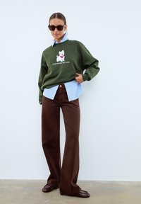 Sweatshirt vert olive avec un graphisme de chien blanc et du texte, superposé sur une chemise rayée bleu clair, associé à un pantalon large marron et des ballerines marron.