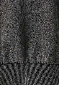 Tejido de sudadera gris oscuro con una textura suave, que presenta un dobladillo de canalé elástico en la parte inferior. Las motas sutiles añaden dimensión.