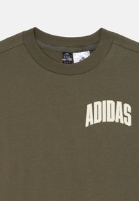 Olive zelená mikina so zúženým okrajom pri krku, s bielym logom "ADIDAS" v ľavom hornom rohu. Materiál vyzerá mäkko a textúrovane.
