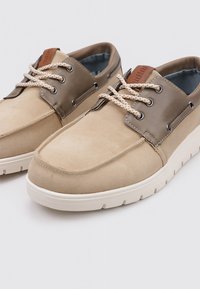 Zapatos de ante beige y piel oliva con suela de goma, punta redondeada, ojales de metal y cordones con patrón. Acento en marrón en el talón.