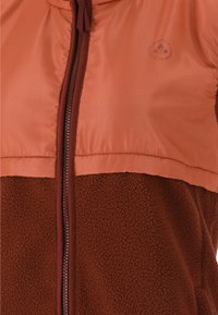 Brun fleecejacka med en orange satin överdel, som har en dragkedja fram och en logotyp på bröstområdet. Mjuk textur över hela jackan.