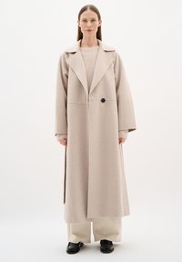 Cappotto in misto lana beige con ampi rever, maniche lunghe e un unico bottone nero. Ha una vestibilità dritta e scende sotto il ginocchio.